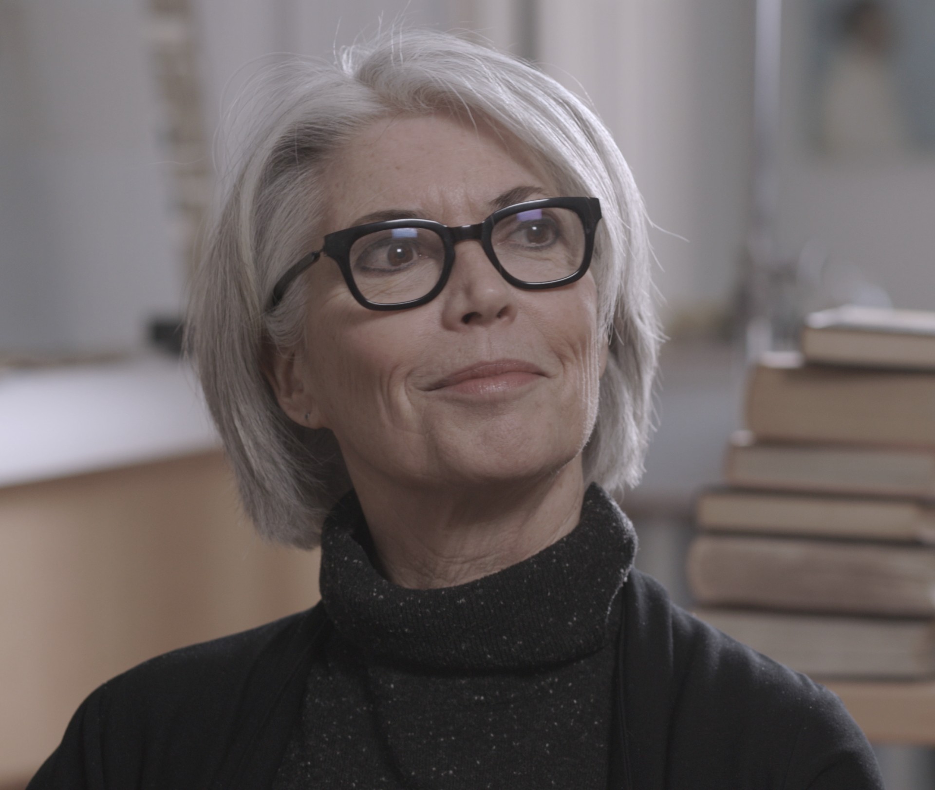 Person with long grey hair, black framed glasses, wearing a black turtle poses for a photo.