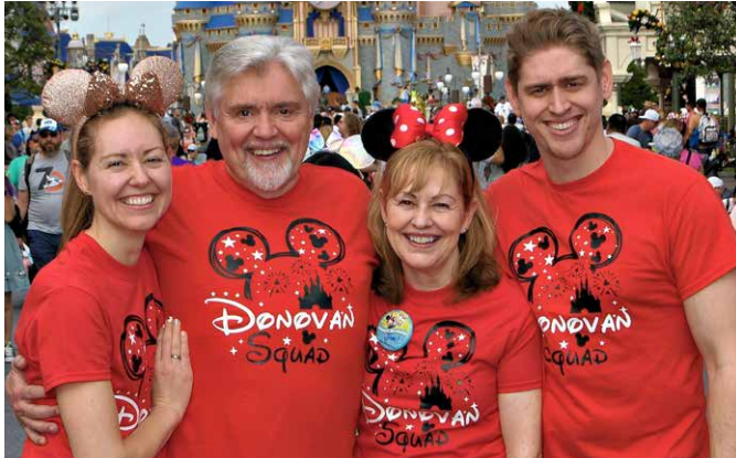A group of four wearing matching red t-shirts with a custom print 'Donovan Squad' posing together at an amusement park, smiling cheerfully with themed headwear.
