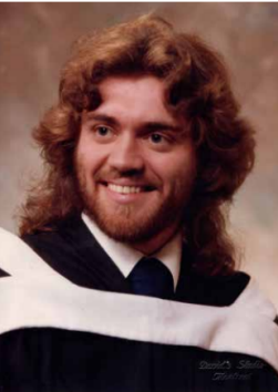 An individual in graduation attire with a smile, featuring dark-colored graduation robes and a white collar, with wavy shoulder-length hair.