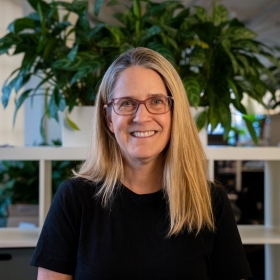 a woman in a casual black shirt smiling in a bright office with plants.