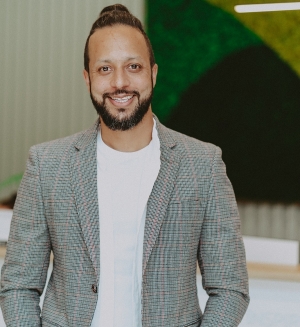 A man wears white t-shirt, an open white button-down, and a grey plaid blazer. He has dark hair, pulled back into a bun, a beard and a mustache. He smiles at the camera. An abstract painting hangs on the wall behind him.