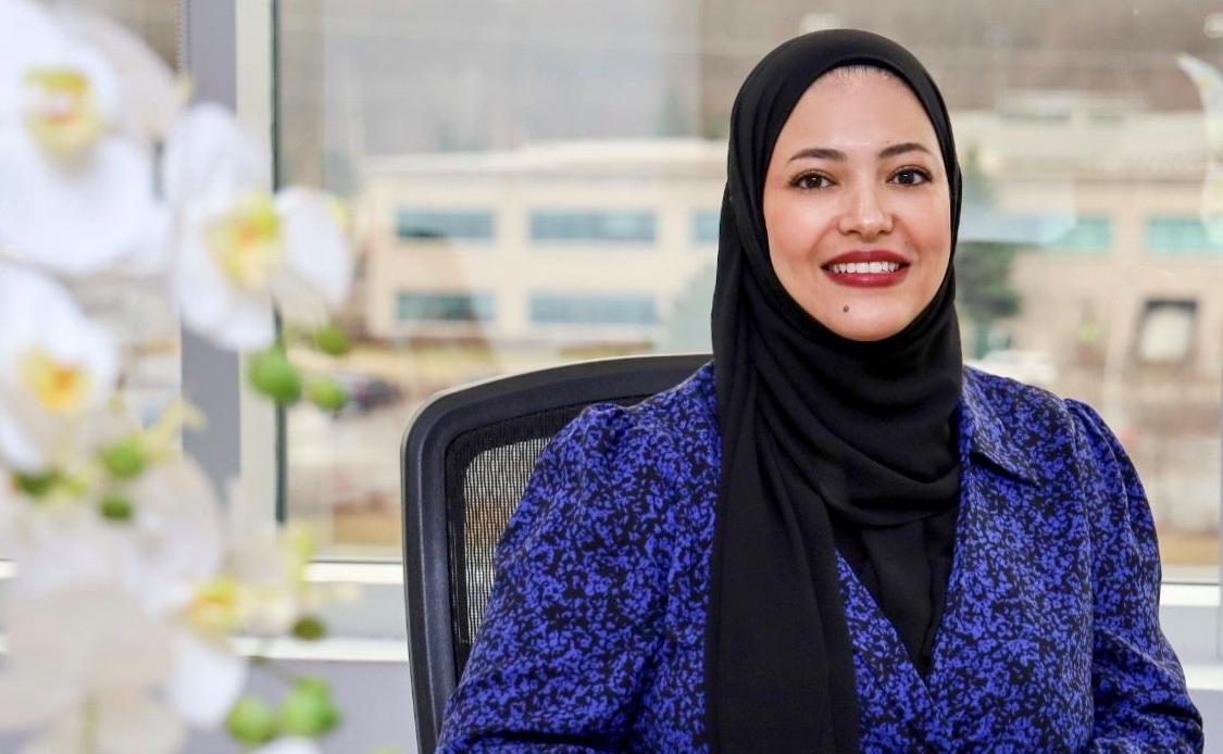 A woman in a professional setting, sitting at a desk with a laptop, smiling confidently.