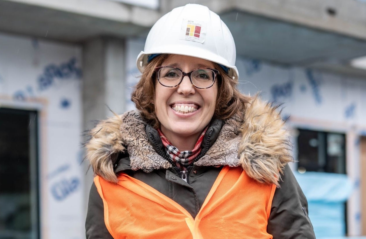 A woman in a construction setting wearing a hard hat and an orange safety vest, smiling confidently.