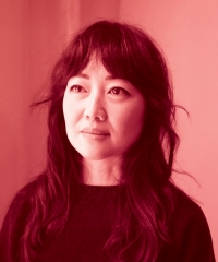Woman with long, dark, wavy hair and a fringe wears a dark, crew-neck top and looks off into the distance