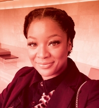 A woman with black hair pulled back into two braids smiles at the camera. She is wearing a black blazer over a leopard-print top with black collar.