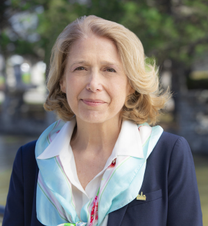 A portrait of a woman with shoulder-length blonde hair, wearing a navy blazer, white blouse, and a light blue scarf with a floral pattern, standing outdoors with trees and water in the background.
