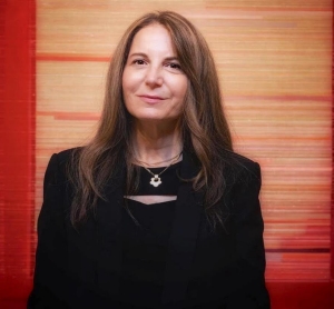 A woman with long brown hair wears a black top with a silver necklace and is standing in front of her artwork, which features horizontal lines in various various shades of red and pale yellow 