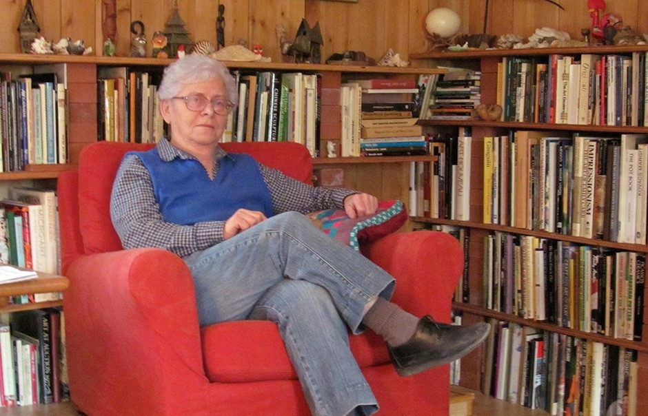 Rose Szasz at home Rose has grey hair and glasses. She sits in a red chair near wooden shelves that are packed with books