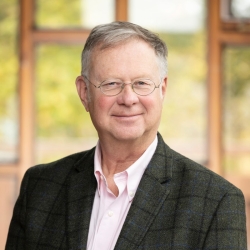 A man in a dark jacket and pink shirt stands in front of a window smiling for the camera