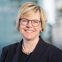 Woman with blonde hair, glasses, wearing a dark jacket smiles for a portrait.