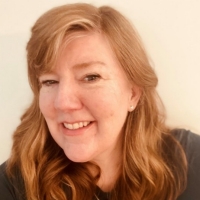 A woman with red hair and pearl earrings smiles for a portrait.