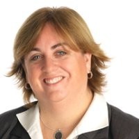 A woman with brown hair, white shirt and a dark jacket sits smiling for a portrait.