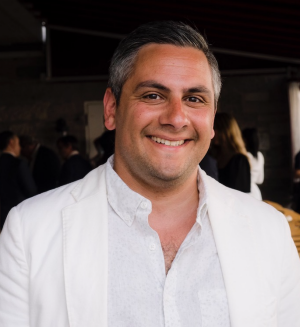 Image of a man smiling. He is wearing a white button-down shirt under a white blazer