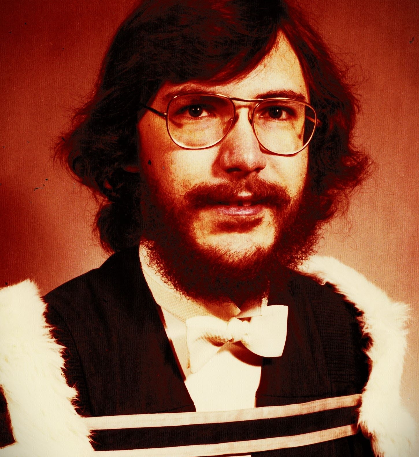 Portrait of a man with long, brown hair, and full beard, wearing glasses and a graduation gown with a white bow tie. 