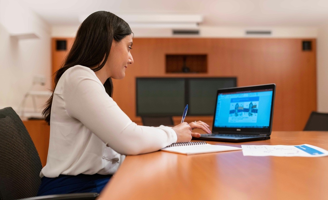 Woman at laptop A woman with dark long hair is wearing a white blouse and sitting at a boardroom table taking notes while looking at a laptop
