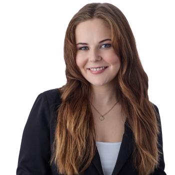 A woman with long brown hair, wearing a white top, blazer and necklace, smiles into the camera.