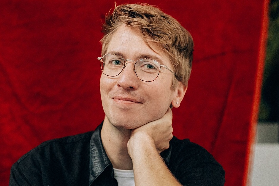Portrait of Devon Bate Devon Bate, who has blonde hair and wire-rimmed glasses, poses in front of a red background, resting his chin on his left hand