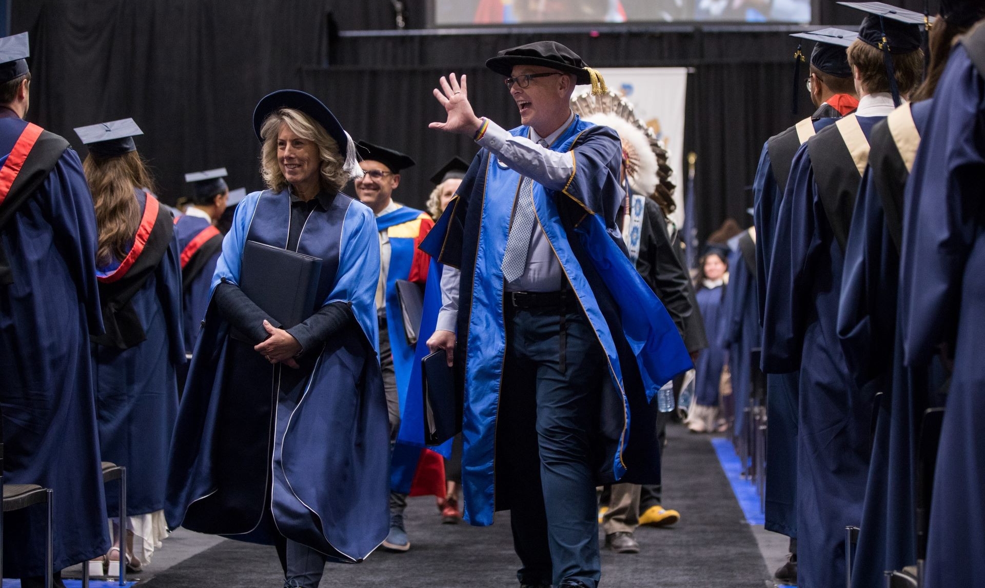 Tim Rahilly is wearing academic regalia, and waves to the audience while walking in a graduation procession alongside other faculty members, surrounded by students in caps and gowns at a convocation ceremony.