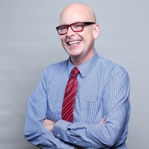 Timothy Rahilly smiles warmly at the camera, dressed in a blue checkered shirt with a red tie, with his arms crossed in front of a plain grey background.