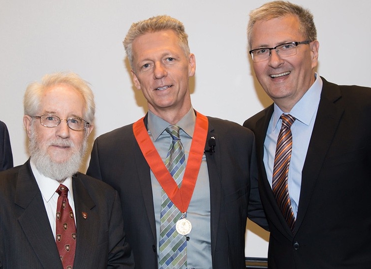 Three men in suits and ties pose for a celebratory photo; the one in the middle has a medal around his neck