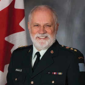 A man with short white hair and white beard smiles with a Canada flag behind him. He is wearing a white shirt and black tie under a Canadian Armed Forces jacket