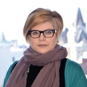A woman with short blonde hair is wearing black-framed glasses and a mauve scarf wrapped around her neck