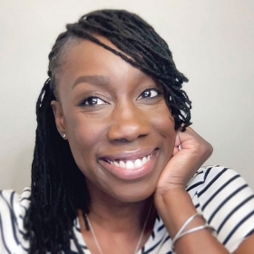 Close up of a woman resting her face on her hand. She is smiling and has long, black hair and is wearing a white shirt with black horizontal stripes