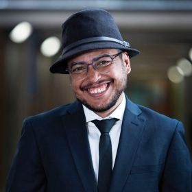 A man with short black beard smiles at the camera. He is wearing a dark blue blazer over a tie and white shirt, and is also wearing glasses and a black fedora