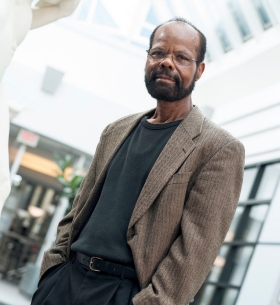 Nigel stands in a sunny atrium. He wears a black T-shirt, black pants and a brown tweed jacket.