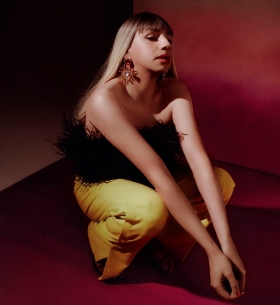 Tranna has long blonde hair. She is posing on a red background, sitting on her heels. She is wearing yellow pants and a furry black tube top. 