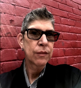 Kat poses in front of a rebrick wall. They have short salt and pepper hair and are wearing dark glasses.