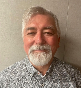 Doug has white hair and a short beard. His cheeks are rosy and he wears a printed grey shirt.