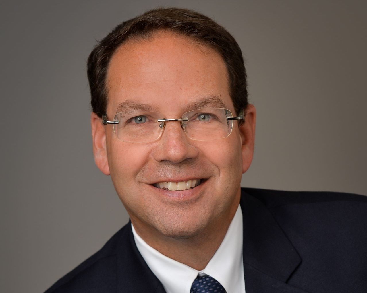 Jon Reider Man in a blue suit, white shirt, blue tie and glasses sit smiling for a portrait.