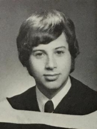 Black and white photo of a young man with dark hair wearing a white shirt and tie with graduation robe 