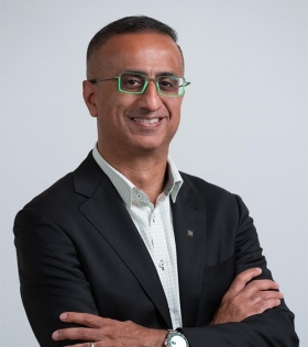 A man with short black hair and glasses smiles at the camera as he stands with his arms crossed. He is wearing a dark blazer over a white button-up shirt