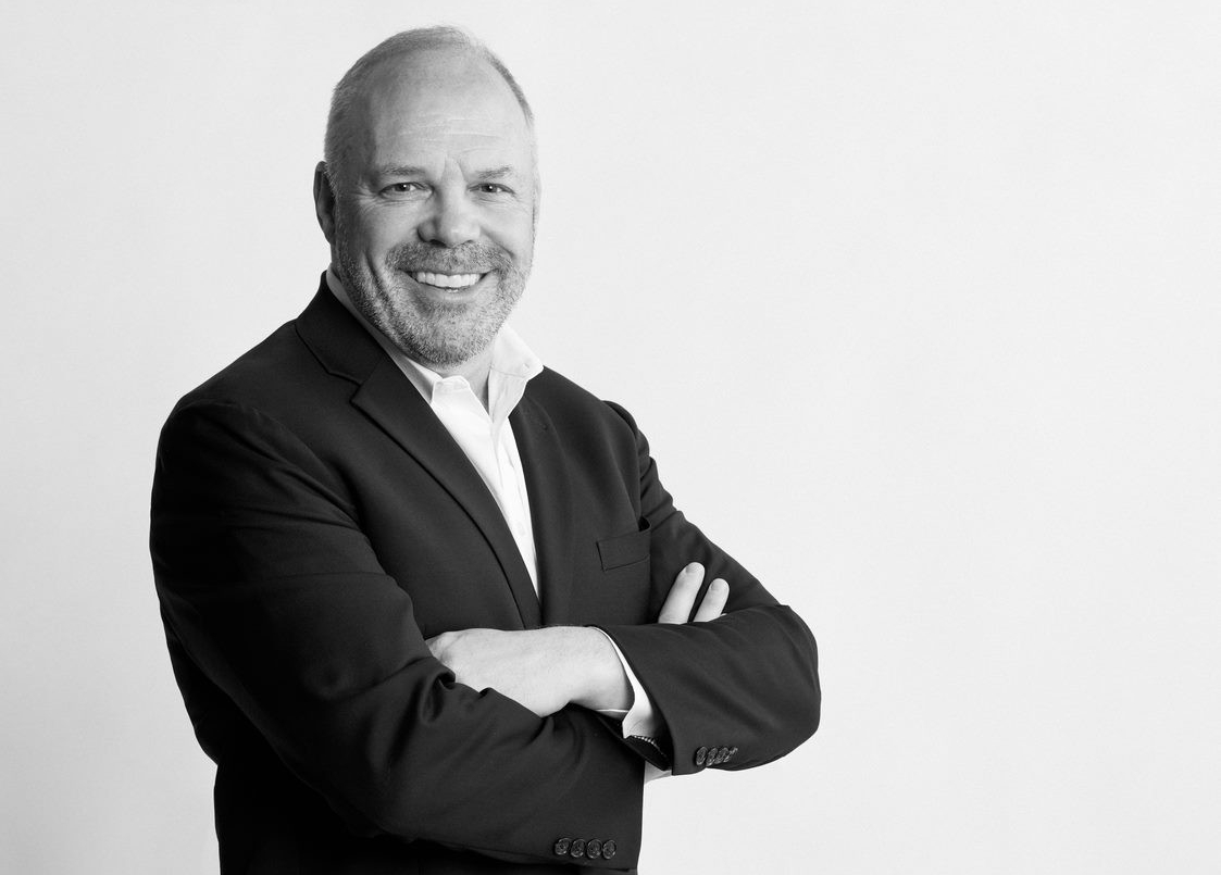 Man in suit jacket and white shirt stands, arms crossed, smiling into the camera