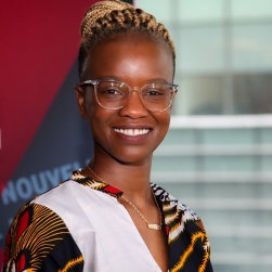 Woman in multi coloured dress and glasses stands in front of a window smiling