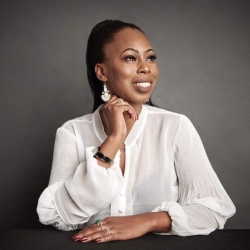A woman with black hair, wearing a white shirt, sits with one arm under her chin and smiles.