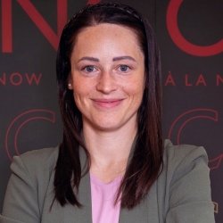 Woman with long brown hair wearing a grey jacket and a pink shirt stands in front of a Concordia backdrop