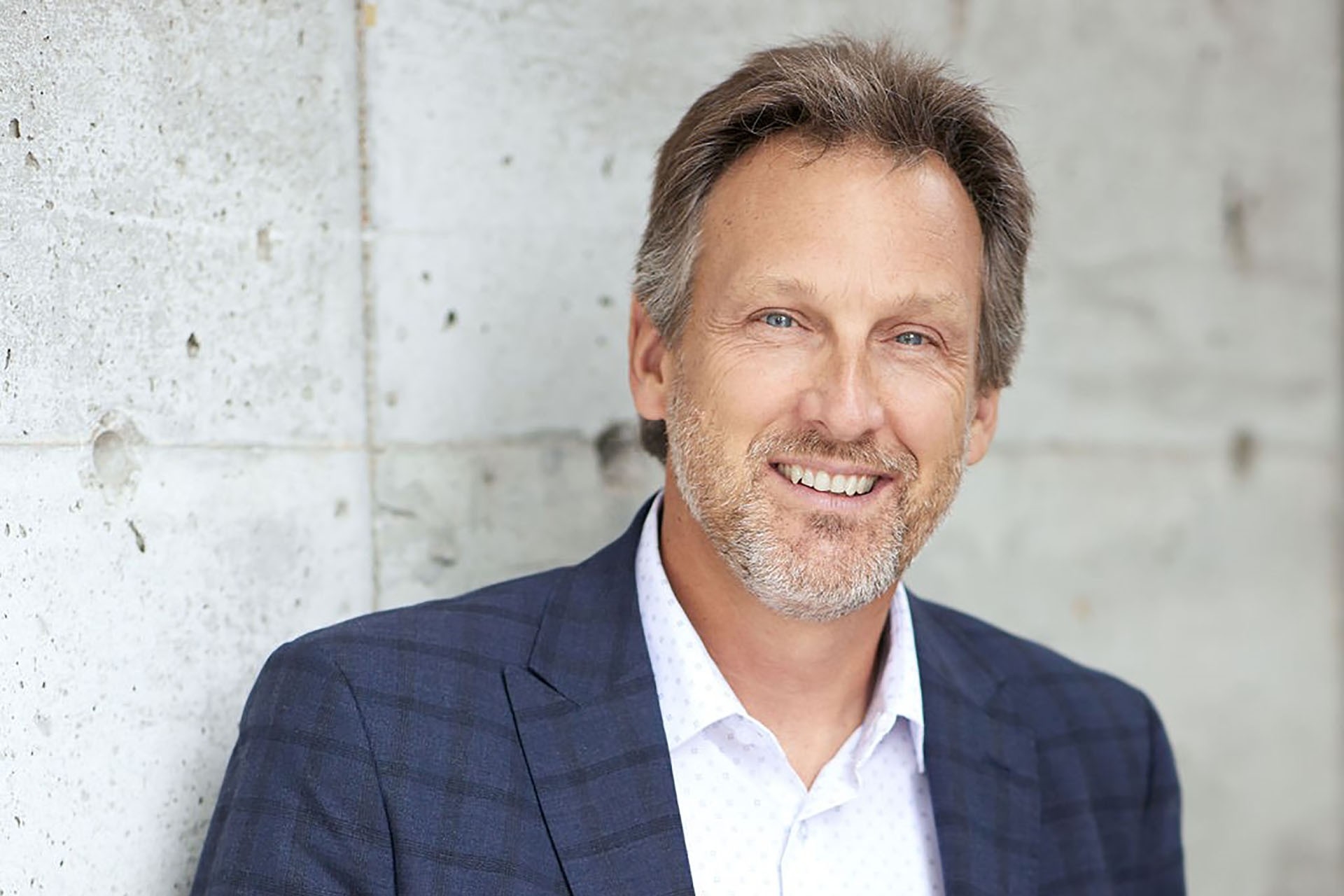 Michel has short gery and brown hair and stands in front of a cement wall. He is wearing a white shirt and a blue blazer. 