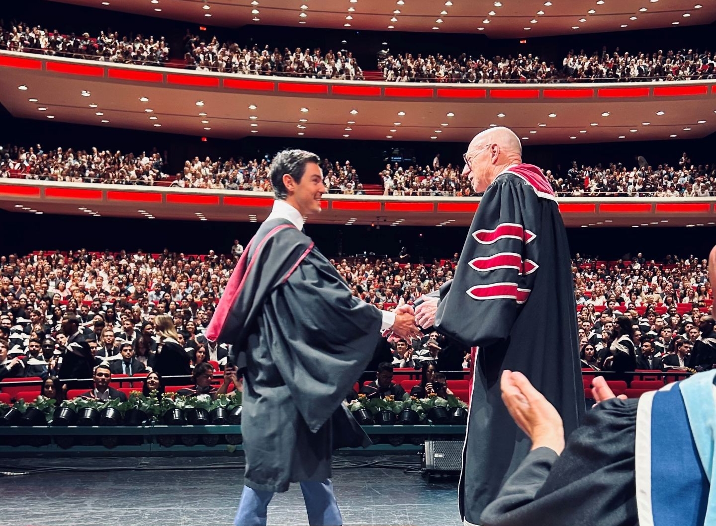 A male student wearing his graduation gown, shakes hands with a university official on stage during his convocation ceremony in a large auditorium filled with attendees.