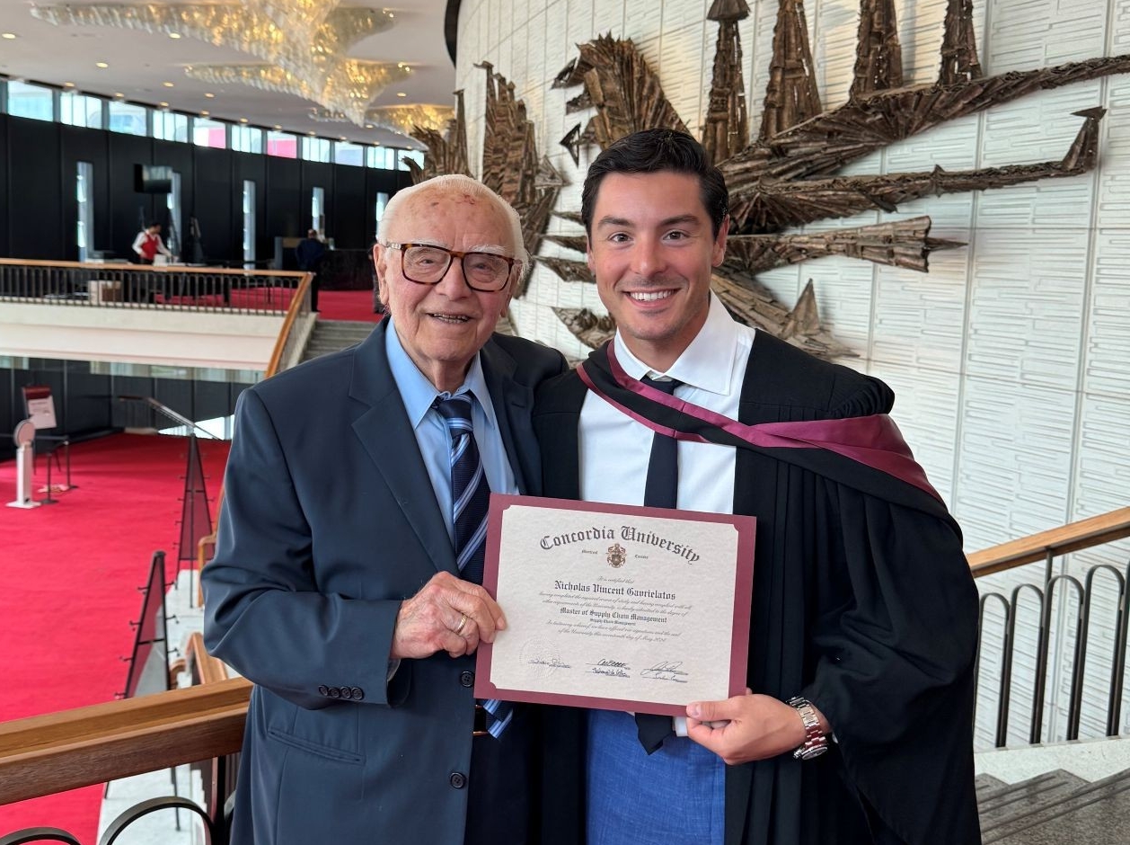 Student in his graduation gown, poses with his grandfather holding his Concordia University diploma in a brightly lit venue with red carpeting and modern decor.