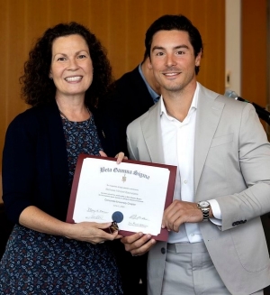 Student dressed in a beige suit, receives a framed certificate from a smiling woman during a Beta Gamma Sigma induction ceremony, with event banners visible in the background.