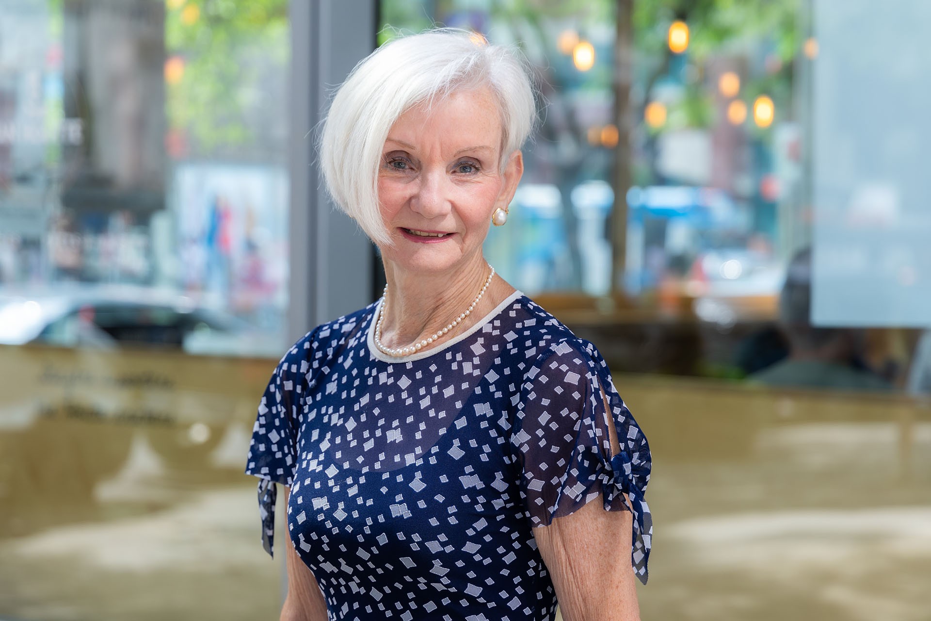 Pamela has white styled into a short, asymmetrical bob. She is wearing a blue dress with a pattern of light blue squares.