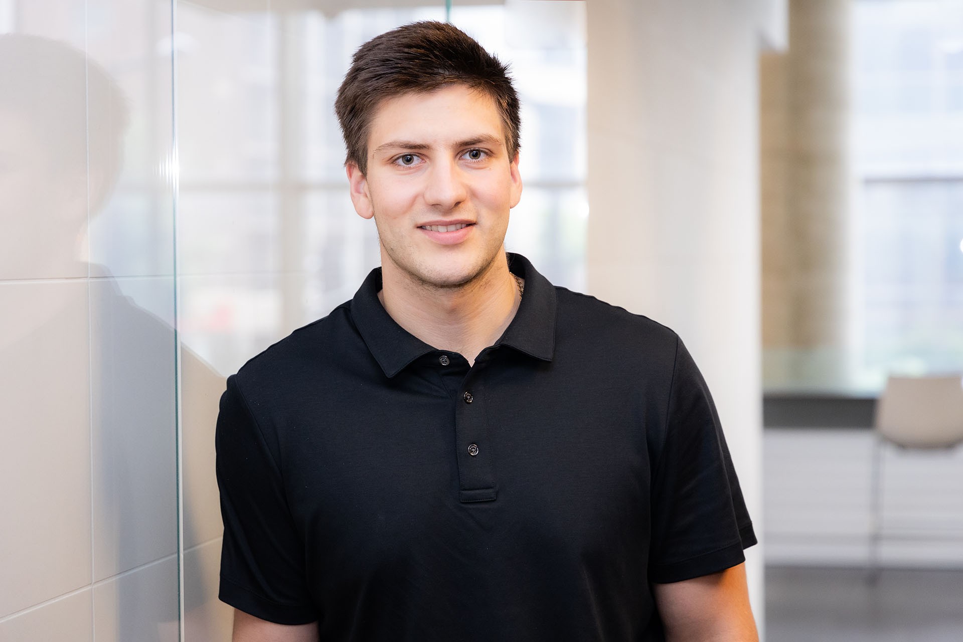 Marcus has short brown hair and is wearing a black short-sleeve, collared T-shirt.