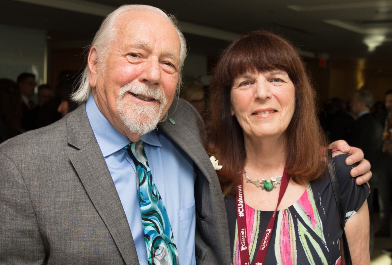 A man and woman stand next to each other and are smiling. The man has his arm around her shoulder