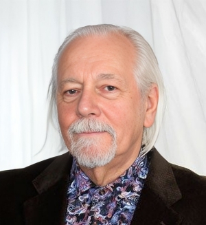 A man with white hair and white goatee faces the camera and is wearing a dark blazer over a paisley shirt