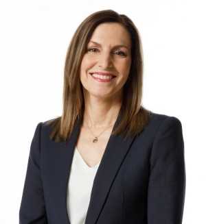 A woman with shoulder-length brown hair is wearing a navy blazer and white blouse. She is standing in front of a white backdrop, smiling