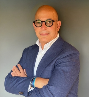 alt="Morgan International's CEO Fadi Ganni wears stands with his arms crossed in front of a grey background. He is wearing dark-framed glasses and a white collared shirt under a blue blazer."