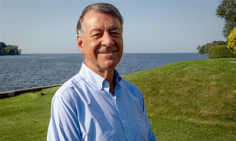 Constantine is standing by a lake with blue water and green grass behind him.